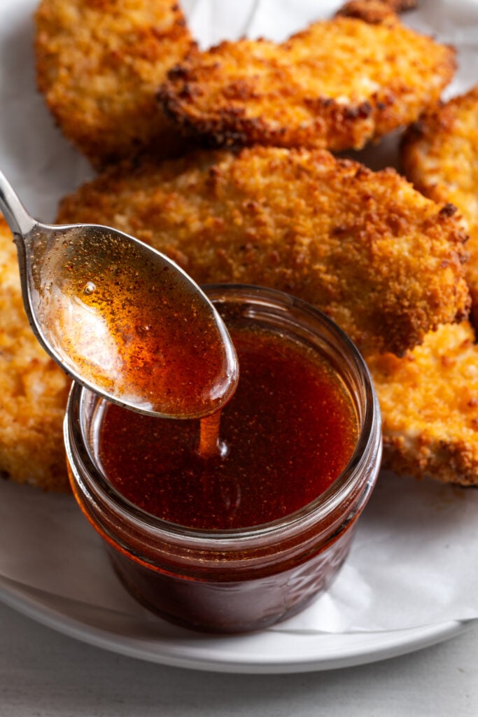 action shot of a silver spoon drizzling homemade hot honey sauce into a small, squat mason jar on a white plate with a pile of crispy air fried chicken tenders.