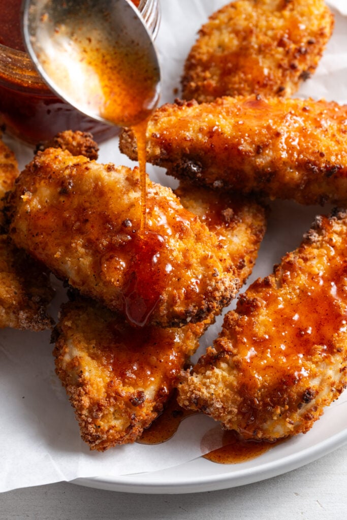 action shot of homemade hot honey sauce being drizzled over spicy air fryer chicken tenders on a white plate.