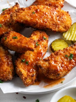 pile of air fryer chicken tenders drizzled in homemade hot honey sauce and sprinkled with parsley, served with a side of sliced pickles in a small ramekin.