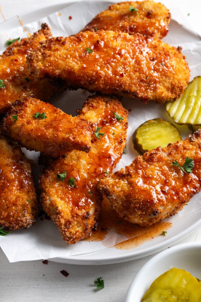 pile of air fryer chicken tenders drizzled in homemade hot honey sauce and sprinkled with parsley, served with a side of sliced pickles in a small ramekin.