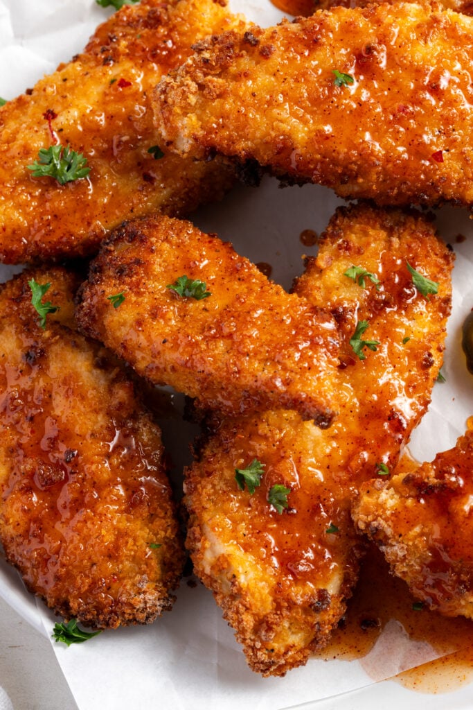 closeup shot of honey hot chicken tenders on a piece of parchment; they're deeply golden brown and the places that the sauce is touching are glistening.