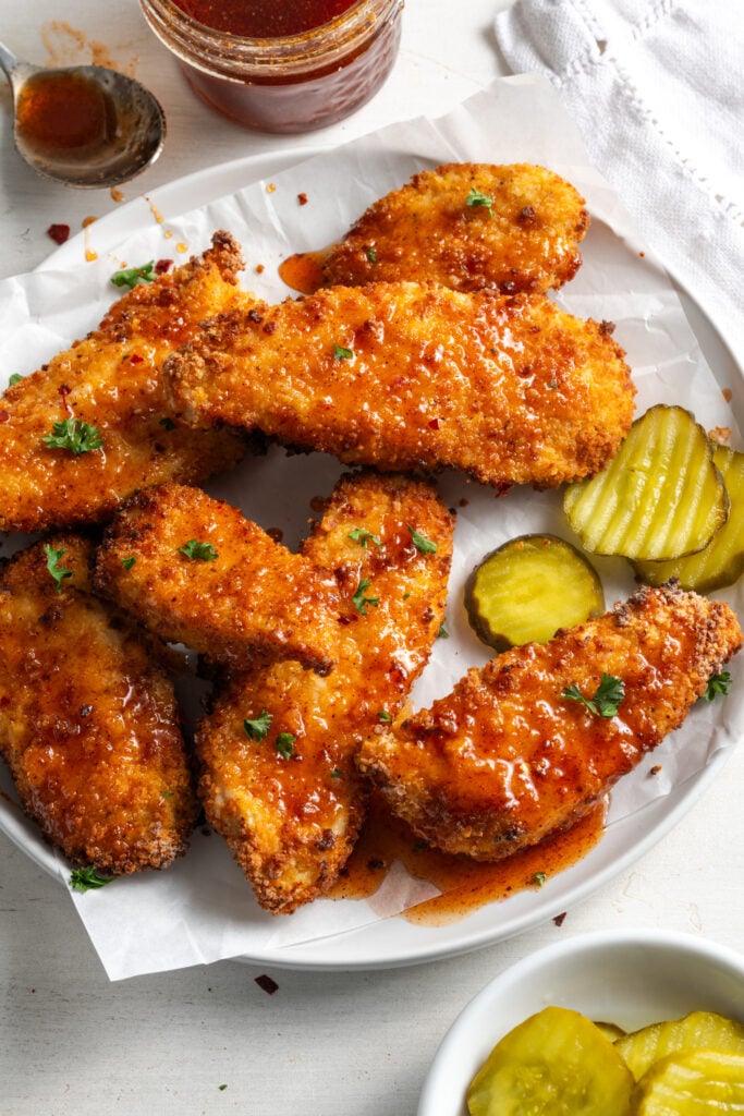 round white dinner plate topped with a crumpled piece of white parchment paper with honey hot chicken tenders drizzled with hot honey sauce, sprinkled with parsley, and served with sliced dill pickles with extra sauce and extra pickle slices in small bowls in the top and bottom of the frame.