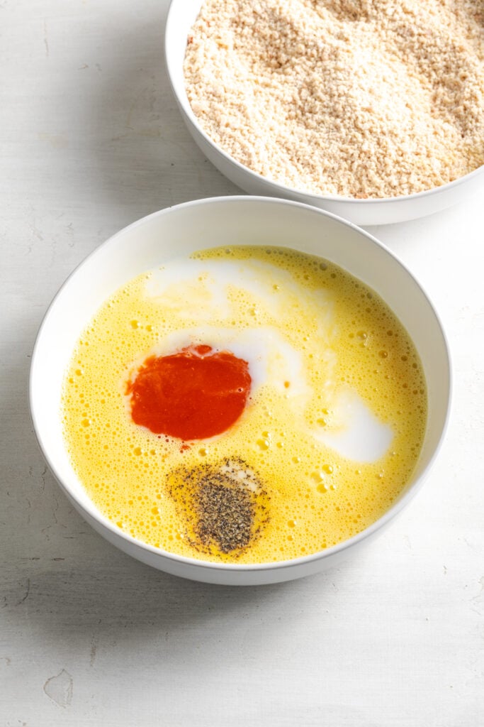 whipped eggs in a bowl with milk, hot sauce, and seasoning added in the center of the frame and the breadcrumb mixture in a separate bowl in the top of the frame.