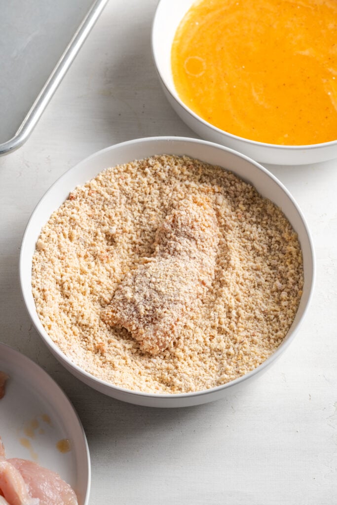 dredging station set up with a single chicken tender that had already been dipped into the egg wash bowl at the top of the frame covered in the breadcrumb mixture in the middle of the frame with a plate of raw, naked tenders in the very bottom corner of the frame.