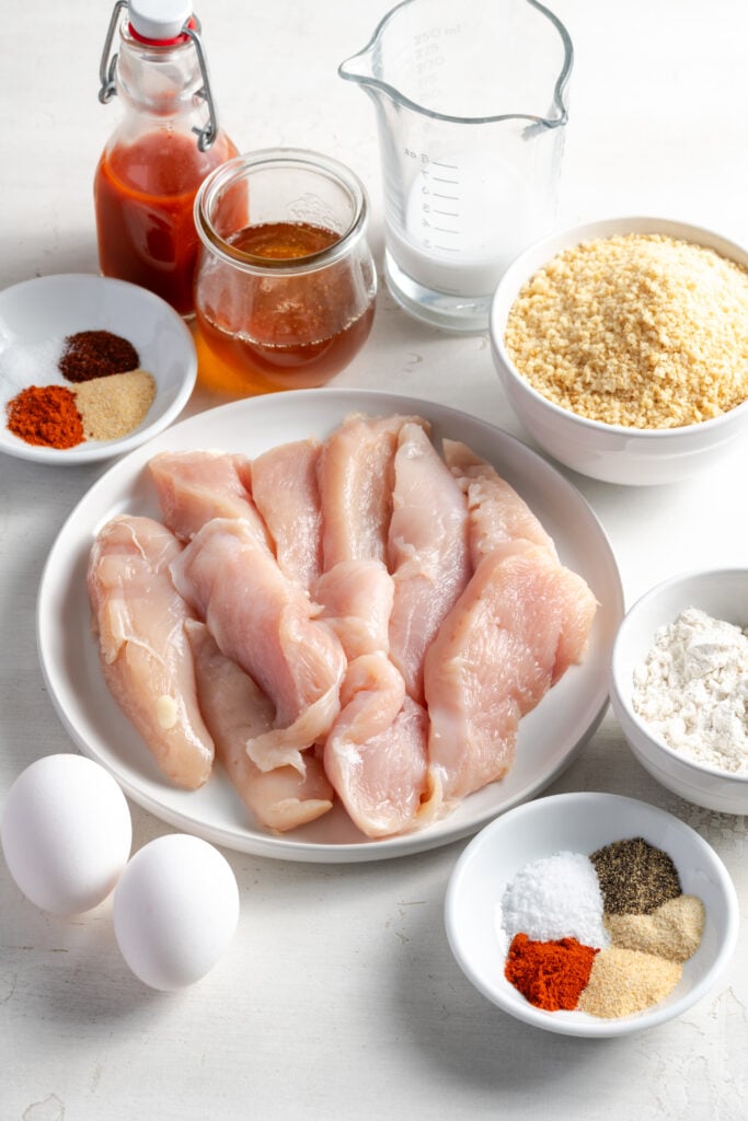 ingredients needed to make hot honey chicken tenders measured out into bowls and plates on a white tabletop.