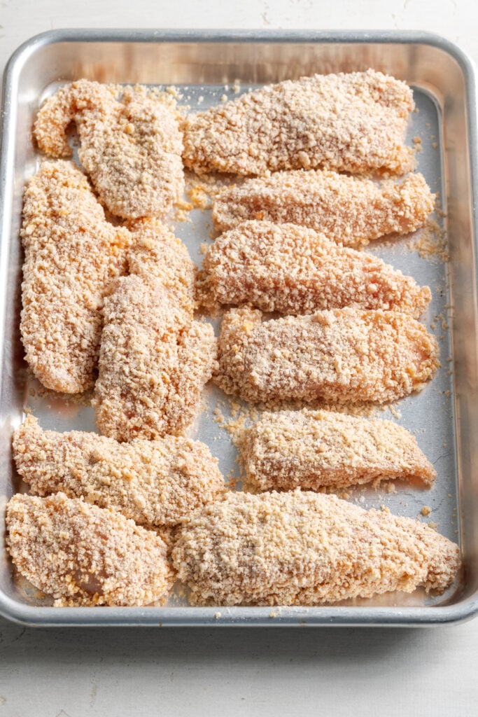 silver baking tray with 12 homemade breaded chicken tenders that have yet to be cooked.