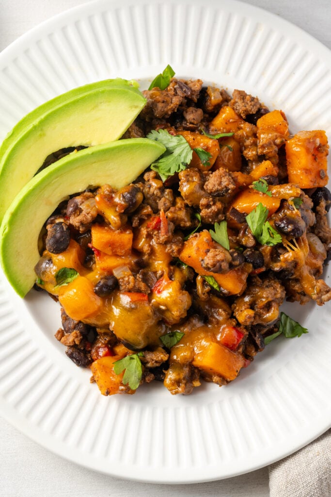 flat lay shot of a round white dinner plate with ribbed edging topped with sweet potato ground beef skillet garnished with cilantro and avocado.