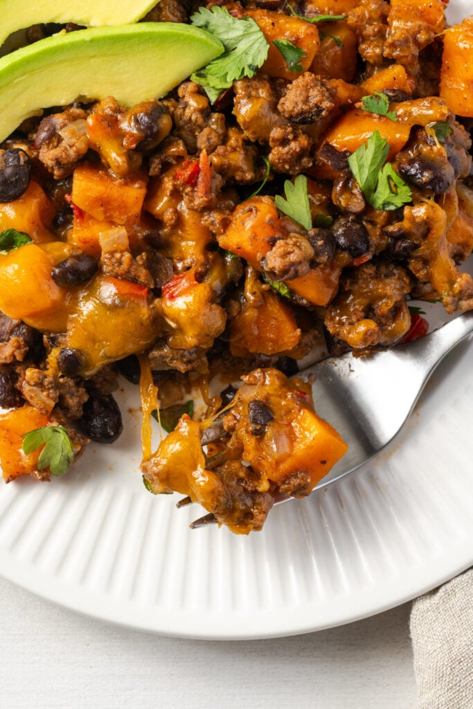 flat lay shot of a silver fork with a cheesy bite of a Tex-Mex ground beef sweet potato skillet on a white plate.