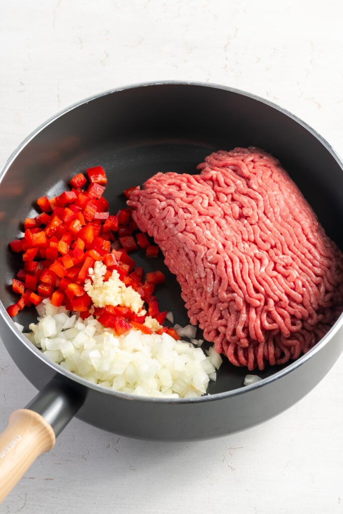 raw ground beef, diced red bell peppers, diced onion, and minced garlic added to a skillet with a wooden handle.