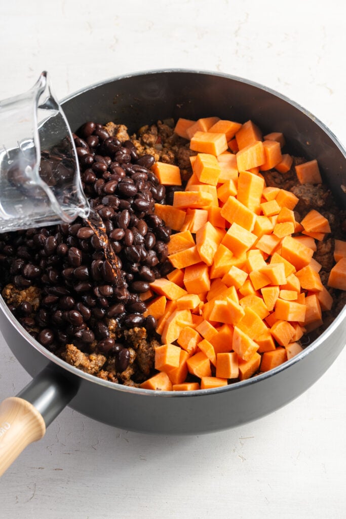 action shot of water being poured into the skillet with the black bean, sweet potato, and beef mixture.