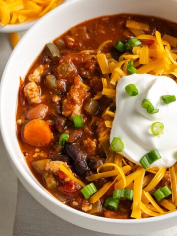 45 degree angle shot of a bowl of red chili with ground chicken topped with cheddar cheese shreds, sliced scallions, and a dollop of greek yogurt with a bowl of extra cheddar cheese in the top left of the frame.