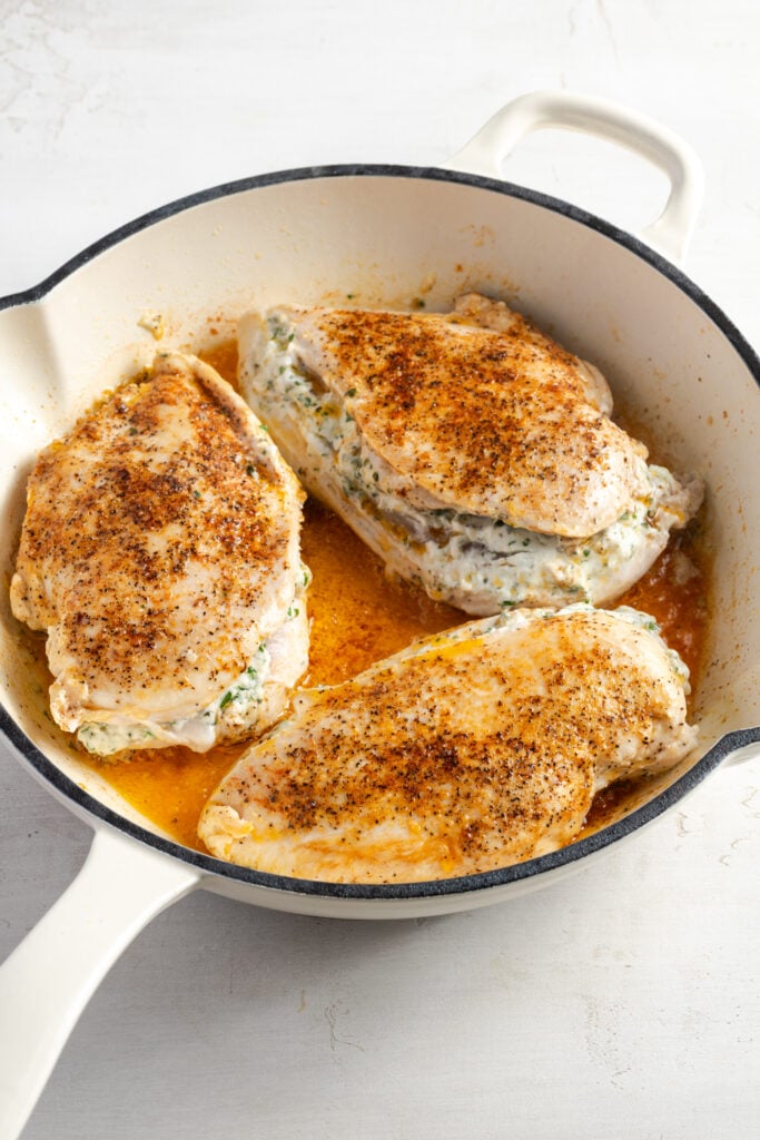 3 seasoned cream cheese stuffed chicken breasts in a white cast iron skillet browning before baking.