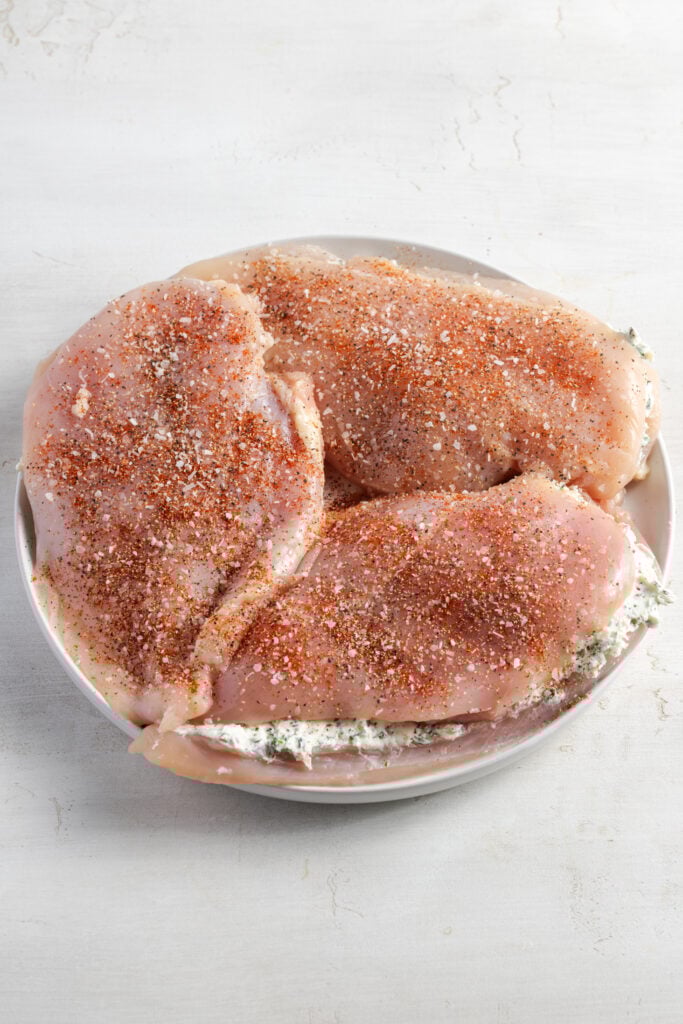 cream cheese stuffed chicken breasts sprinkled with seasoning on a white plate before cooking.