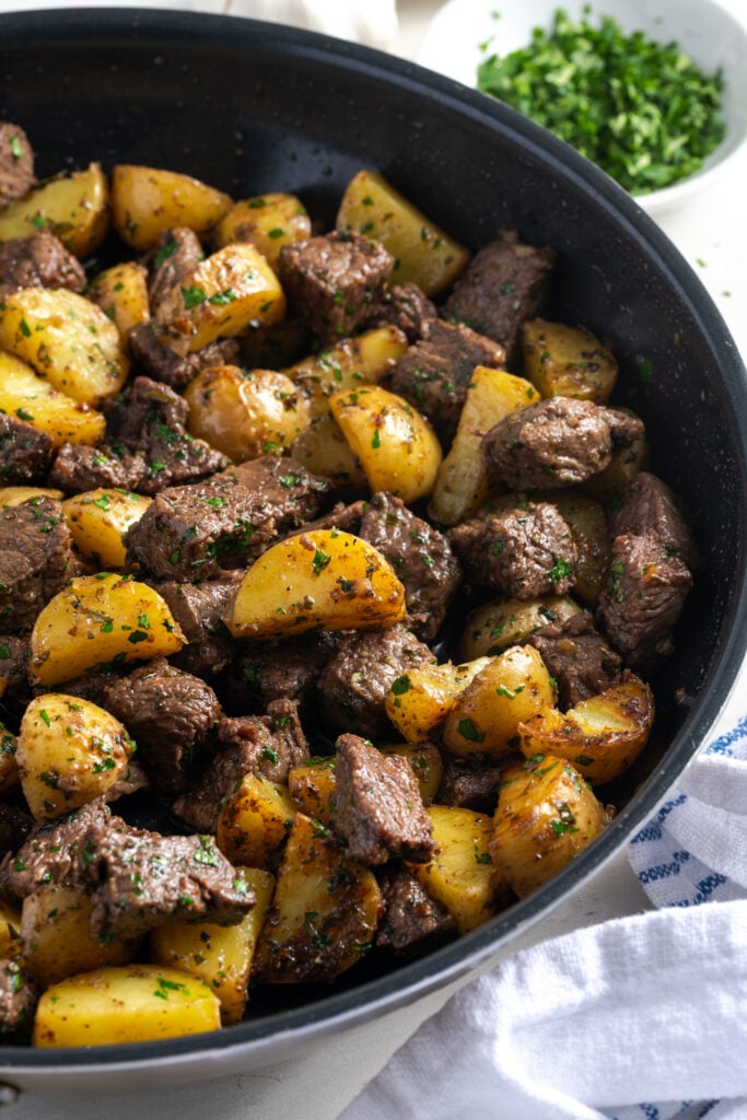 half of a black skillet of garlic steak bites and potatoes on a white table with a small white bowl of chopped fresh parsley.