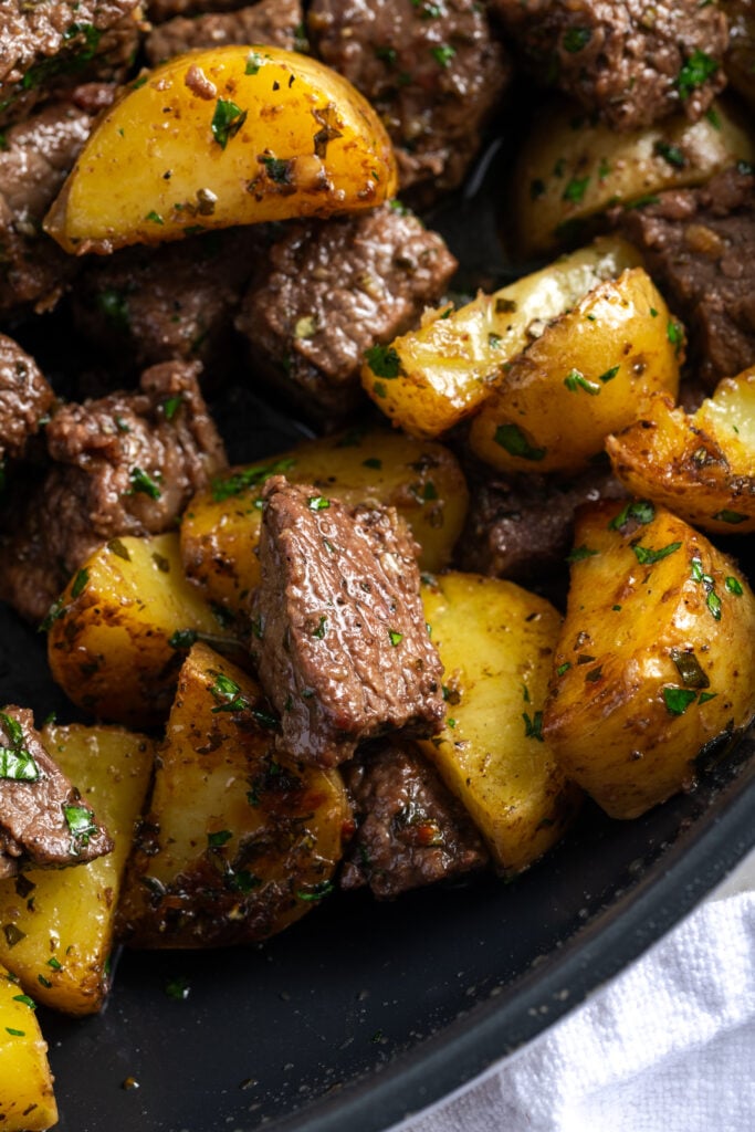 closeup shot of the corner of a black skillet filled with garlic steak bites and potatoes.