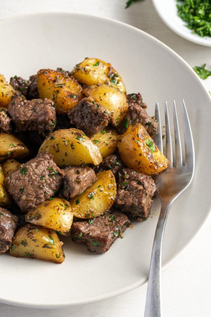 closeup shot on three quarters of a round white plate with garlic and butter steak bites and potatoes with a silver fork in the main frame, with a small bowl of parsley in the top right.