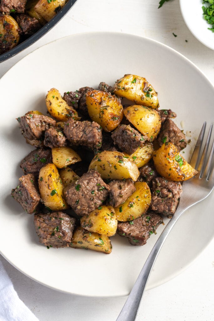 round white dinner plate with juicy steak bites and potatoes in a garlic butter sauce with a silver fork.