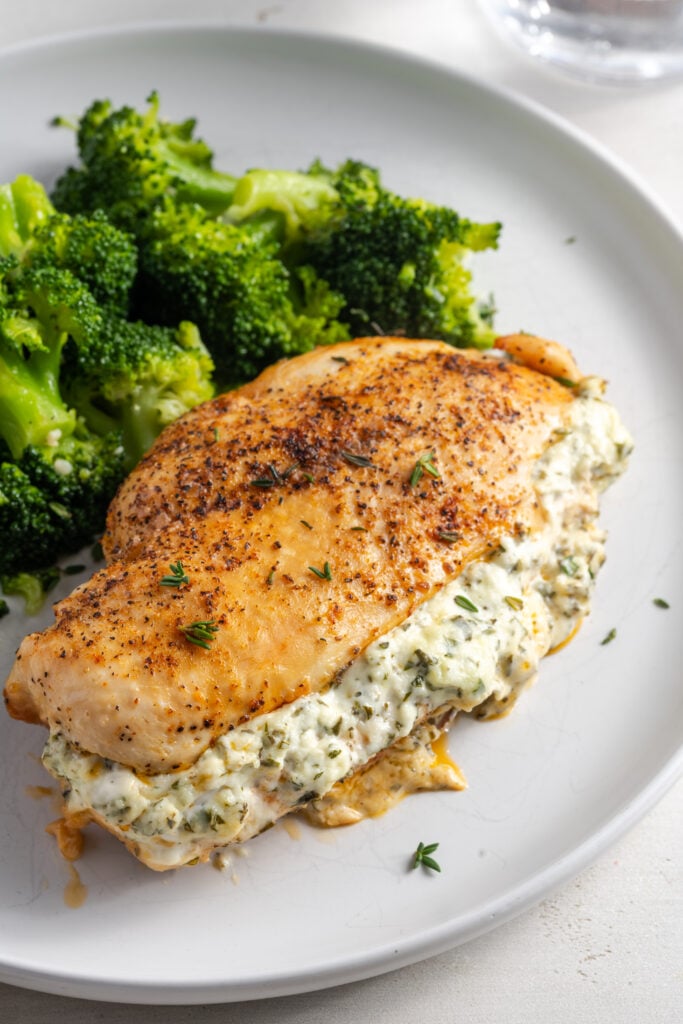 cream cheese stuffed chicken breast on a white dinner plate topped with fresh thyme leaves and a side of broccoli.