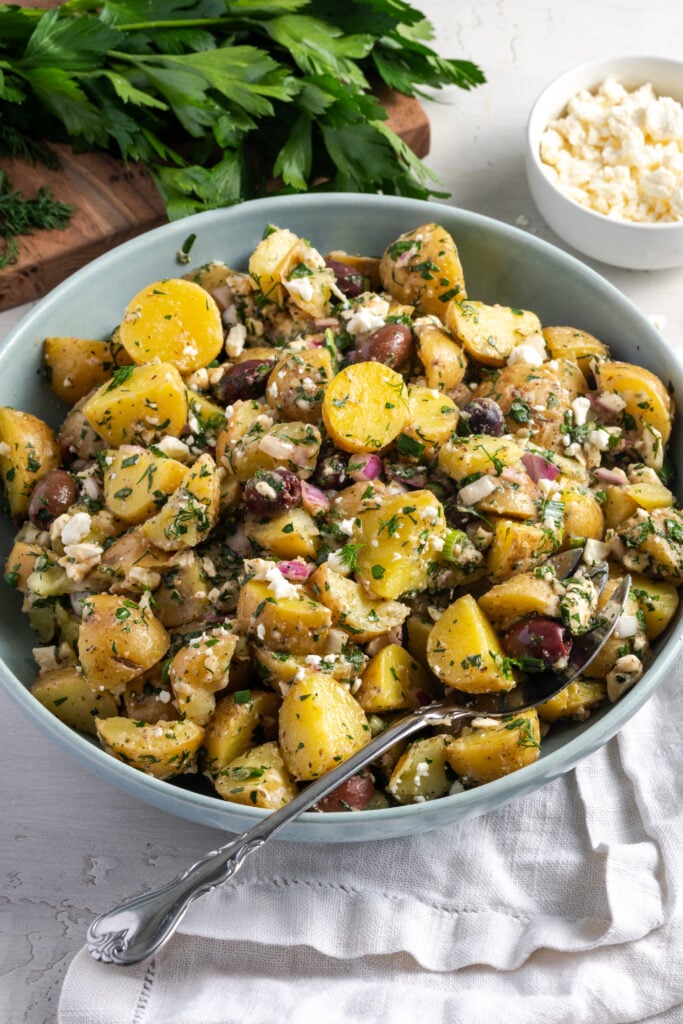 pale blue serving bowl of greek potato salad made without mayo on a white table with a bunch of parsley on a wooden chopping board in the top left of the frame.