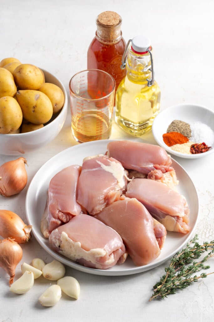 ingredients needed for making chicken thighs and potatoes recipe measured out into white bowls and plates on a white table.