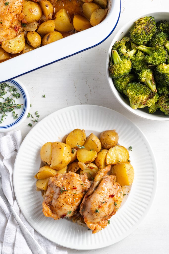 flat lay shot of a white table with a 9x13 baking dish of chicken thighs and potatoes with a white dinner plate with a serving plated out and a bowl or roasted broccoli with some raw thyme in a small pinch bowl and scattered on the table.