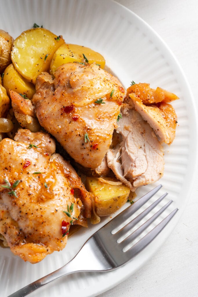 closeup shot of the righthand side of a dinner plate of chicken thighs and potatoes with a few slices of the top chicken thigh rested on the side to show the juicy interior with a silver fork resting on the side of the plate.