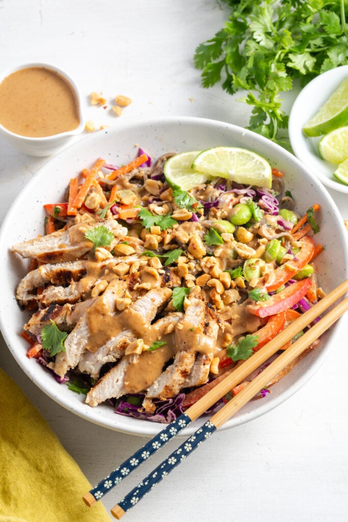 white table with a serving bowl of veggie packed thai peanut noodles with chicken with a side of extra thai peanut salad dressing, a small bowl of sliced limes, a bunch of fresh cilantro and a yellow napkin framing it.