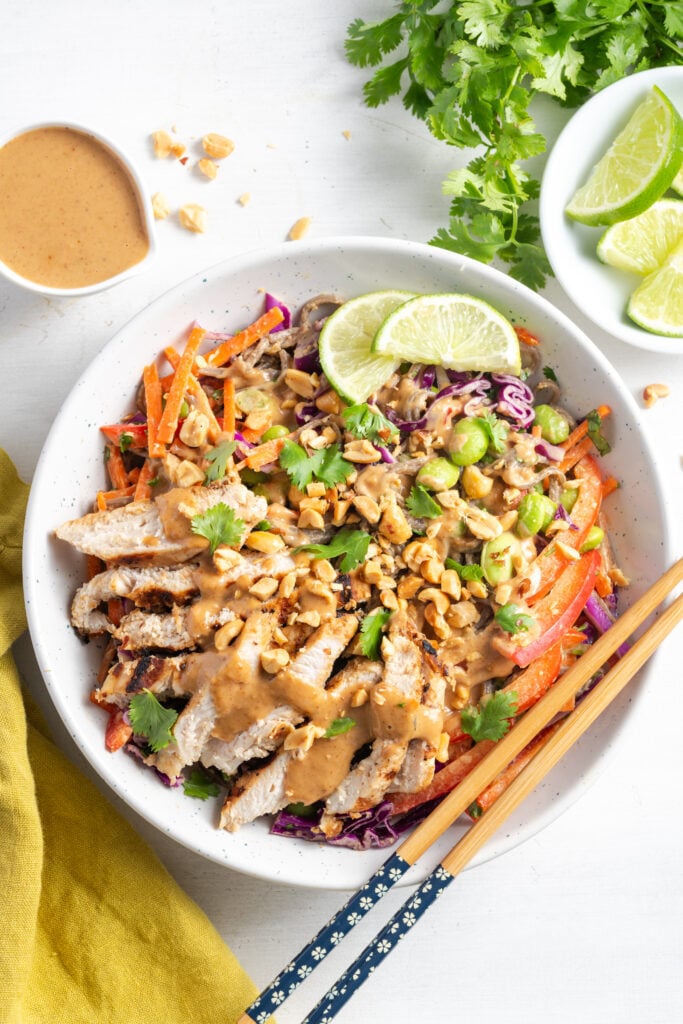 flat lay hero shot of plated thai peanut noodle salad recipe in a white bowl on a white table with extra ingredients scattered around.