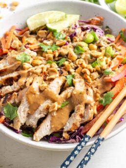 white bowl with Thai peanut noodles and chicken with a pair of wooden chopsticks balancing on the side of the bowl.