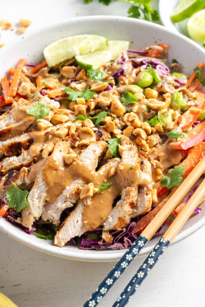 white bowl with Thai peanut noodles and chicken with a pair of wooden chopsticks balancing on the side of the bowl.