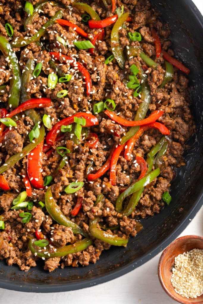 flat lay shot of a skillet of ground beef and pepper strips in an Asian sauce with a small pinch bowl of sesame seeds in the bottom right corner of the frame.