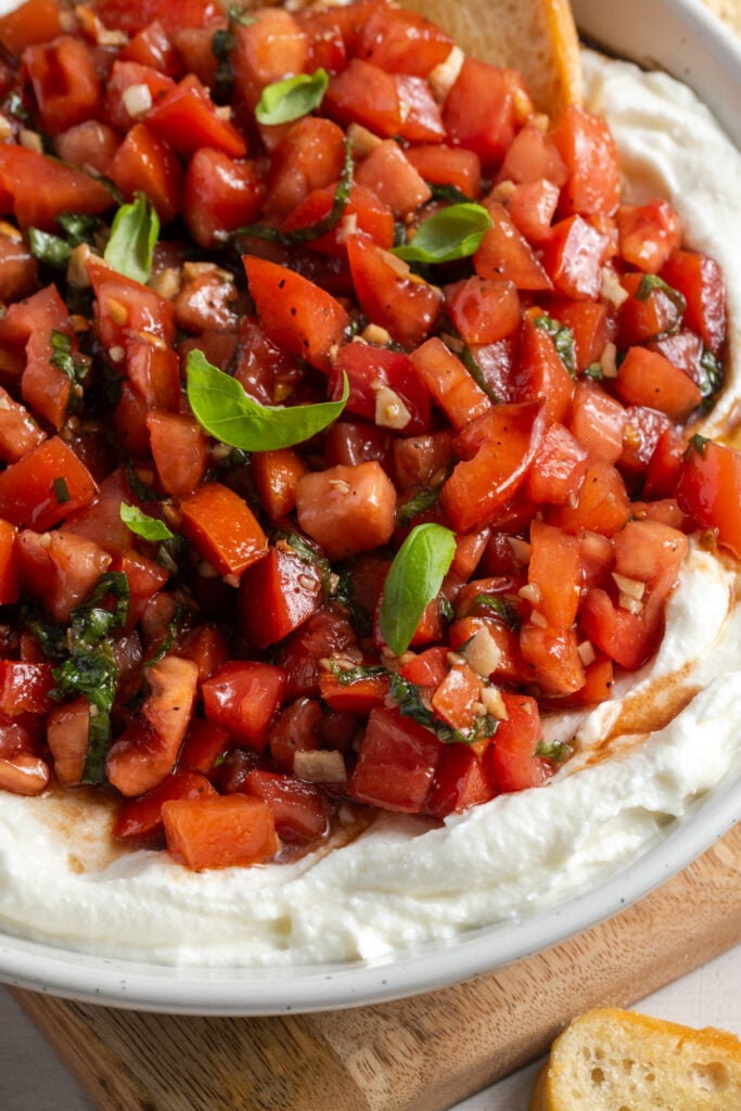 bruschetta appetizer dip in a white serving bowl on a pale wooden cutting board on a white table.