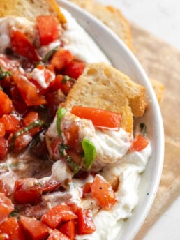 bruschetta dip on a crostini in the serving bowl, showing both the creamy whipped ricotta and the tomato topping.