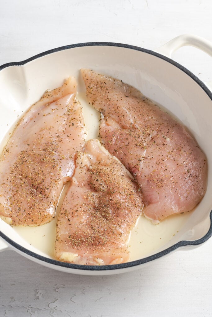 raw seasoned chicken breasts being seared in a white skillet.