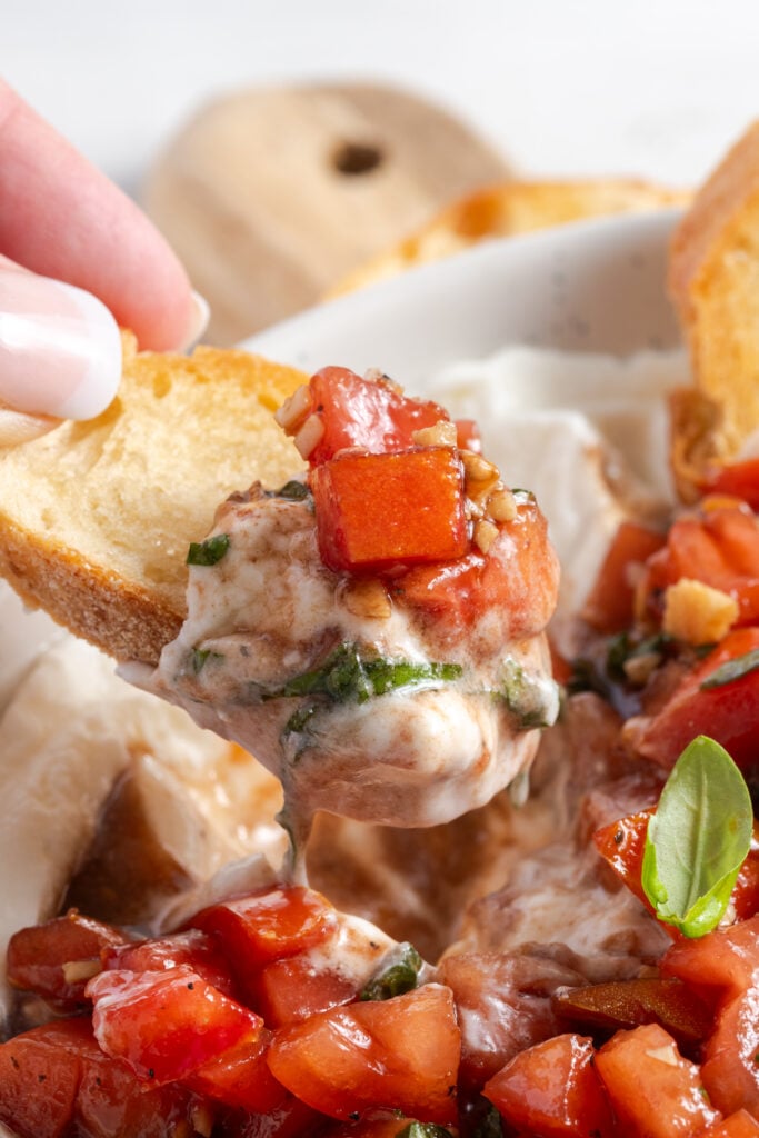 hand dipping a crostini slice into the bruschetta dip, mixing the creamy whipped ricotta with the juicy tomato mixture.