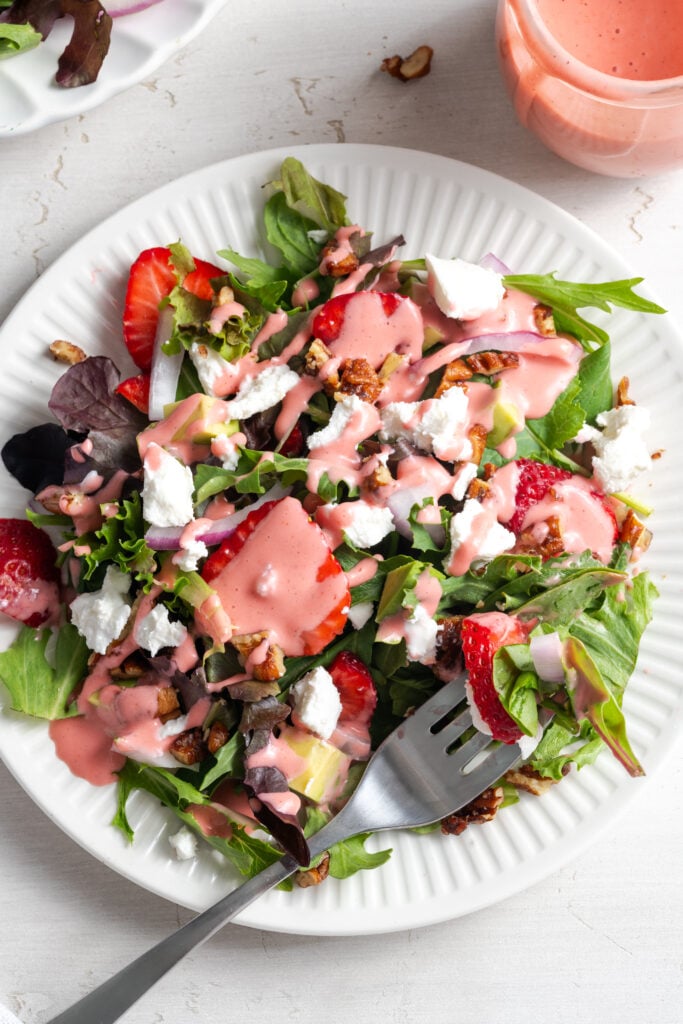 white salad plate with a silver fork taking a bite of goat cheese strawberry salad with candied pecans on a white table with a small jar of extra strawberry dressing.