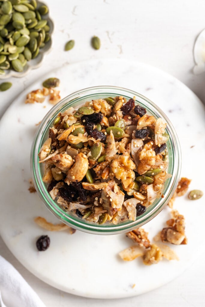 flat lay shot of grain-free granola in a jar on a white marble round serving tray on a white table with some of the granola scattered around.