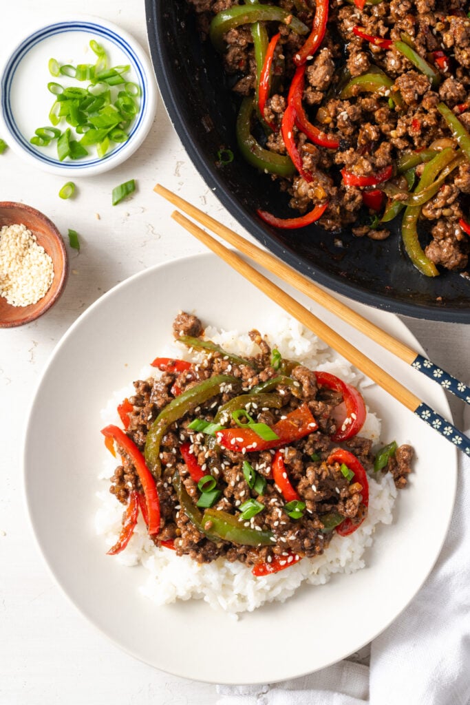 ground beef and peppers over rice with a pair of wooden chopsticks and two small pinch bowls with scallions and sesame seeds and the rest of the skillet with the stir fry.