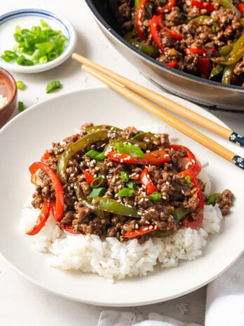 45 degree angle shot of a ground beef pepper rice bowl with wooden chopsticks on a white table with a small bowl of scallions and the rest of the skillet with the stir fry.