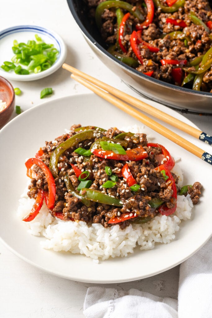 45 degree angle shot of a ground beef pepper rice bowl with wooden chopsticks on a white table with a small bowl of scallions and the rest of the skillet with the stir fry.