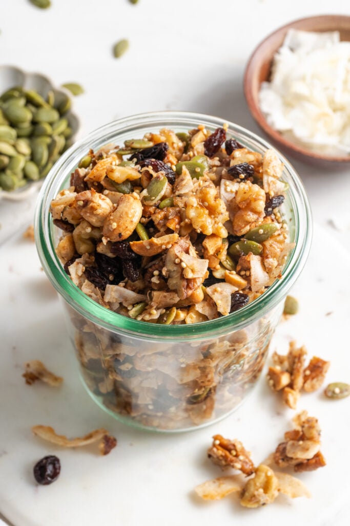 nut and seed grain free granola in a clear french mason jar with two smaller bowls of pepitas and unsweetened coconut flakes on the white table and some of the granola and seeds scattered around.