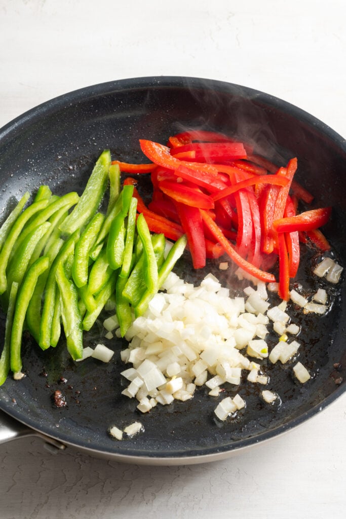 diced onions and sliced bell peppers (red and green) added to a hot skillet with oil.