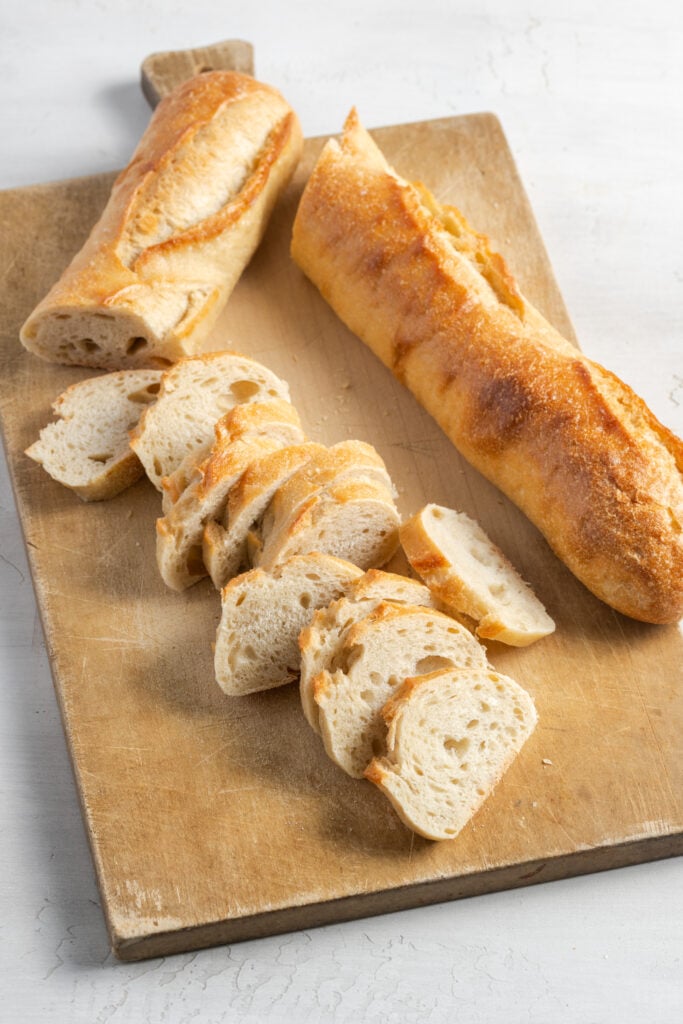 baguette on a wooden cutting board cut in half; half of one half has been sliced into crostini shape.