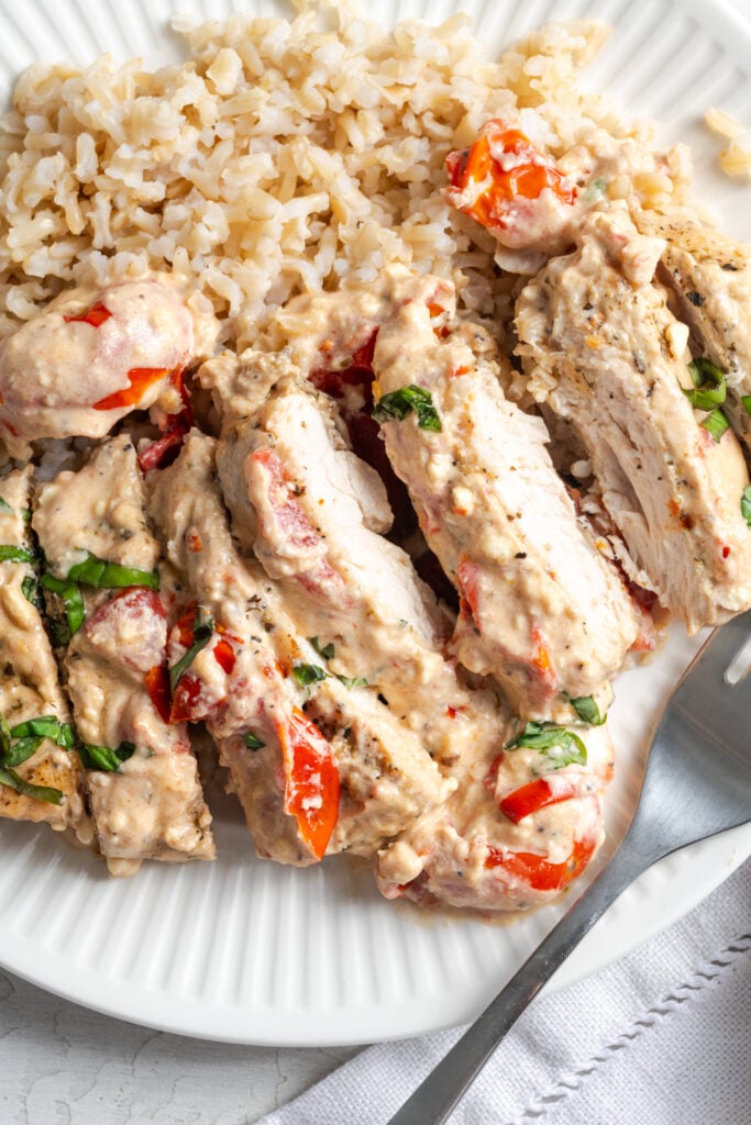 sliced feta tomato baked chicken on a white dinner plate with brown rice and a silver fork.