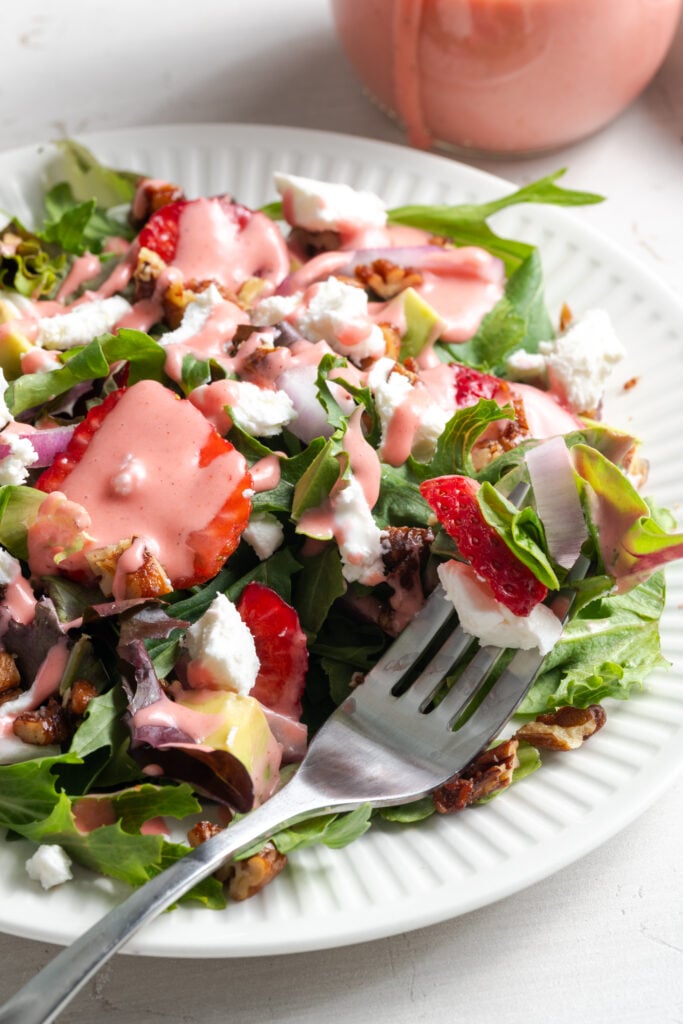 45 degree angle shot of a silver fork on a plate with a bite of goat cheese strawberry salad with strawberry dressing, candied pecans, and fresh greens.
