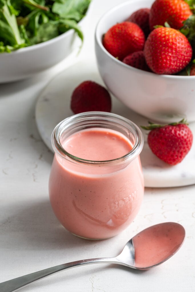 closeup shot of a small glass jar of homemade strawberry vinaigrette on a white table in front of a bowl of fresh strawberries and another bowl of mixed greens.