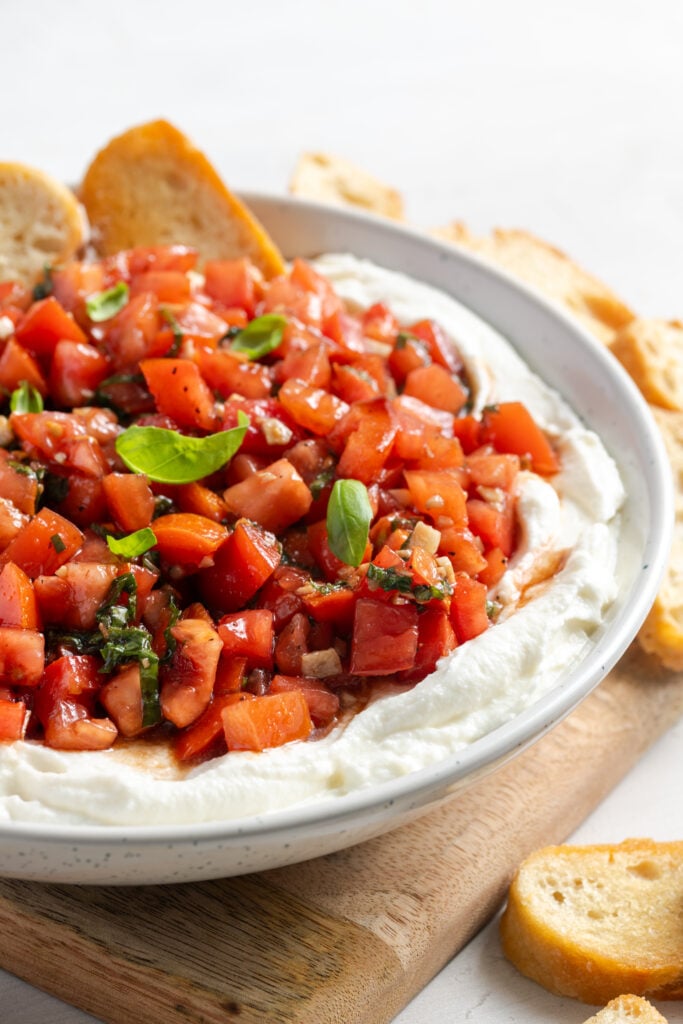 closeup shot of the right half of a bowl of whipped ricotta bruschetta dip topped with fresh basil leaves and a few crostini sticking out the side.