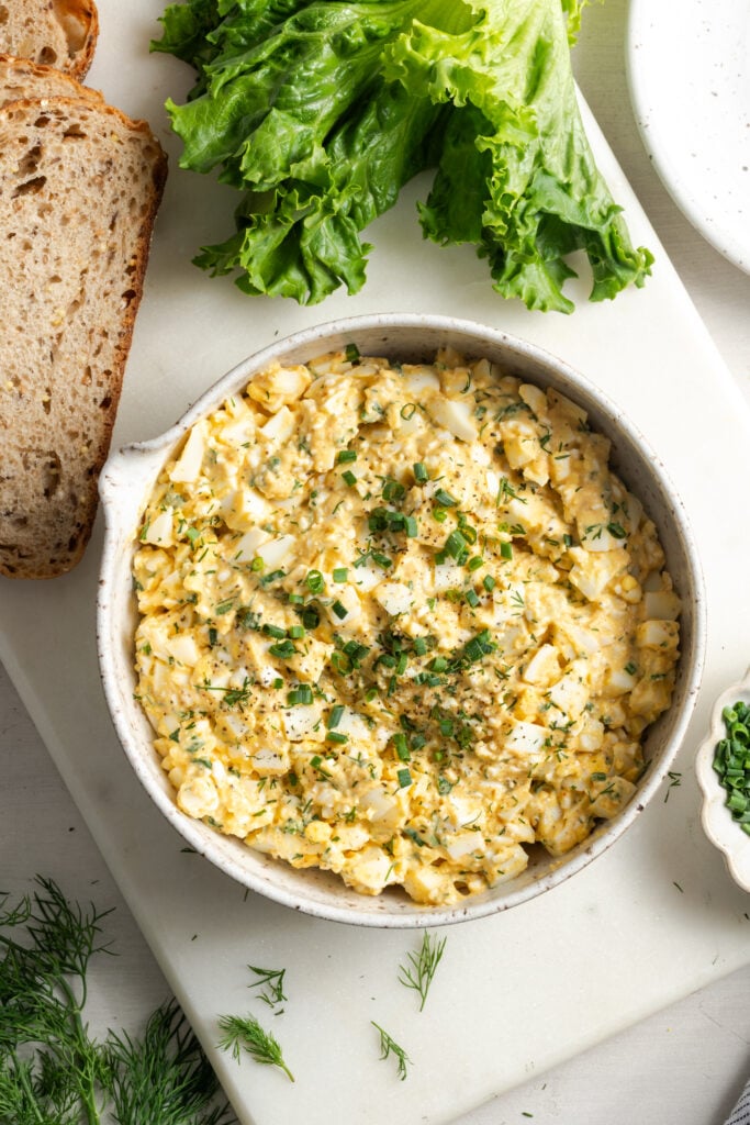 flat lay shot of a bowl of cottage cheese and egg salad on a white cutting board with sliced multigrain bread and a head of leaf lettuce.