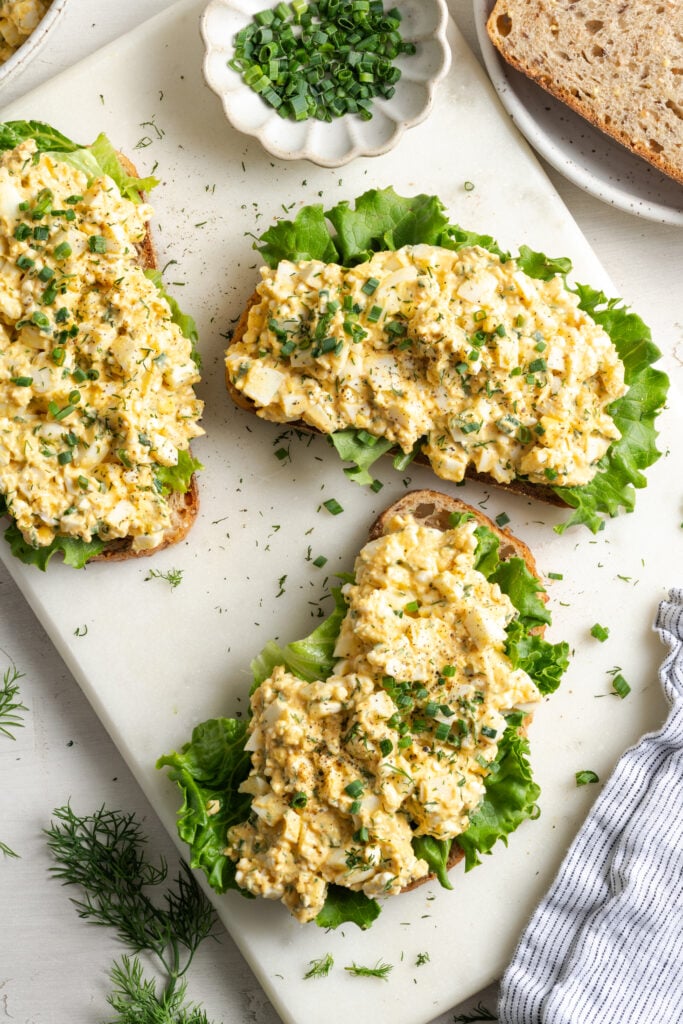 flat lay shot of 3 pieces of toast topped with fresh leaf lettuce and the cottage cheese egg salad mixture, garnished with fresh herbs from a pinch bowl nearby.