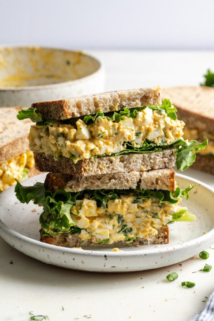 halved and stacked cottage cheese-based high-protein egg salad sandwich on a white plate on a slightly messy white work surface with more sandwiches in the background and a few pieces of fresh herbs scattered around.